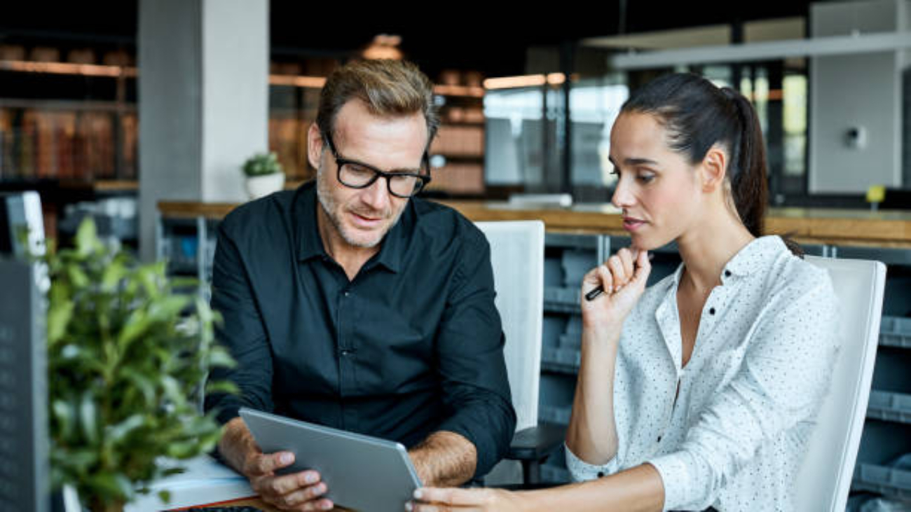 Male and female colleagues looking at tablet PC. Business people are working at desk. They are sitting in textile factory.